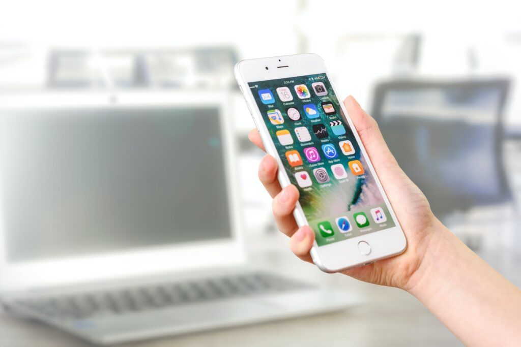 hand holding an iPhone with apps in front of a blurred background of a computer