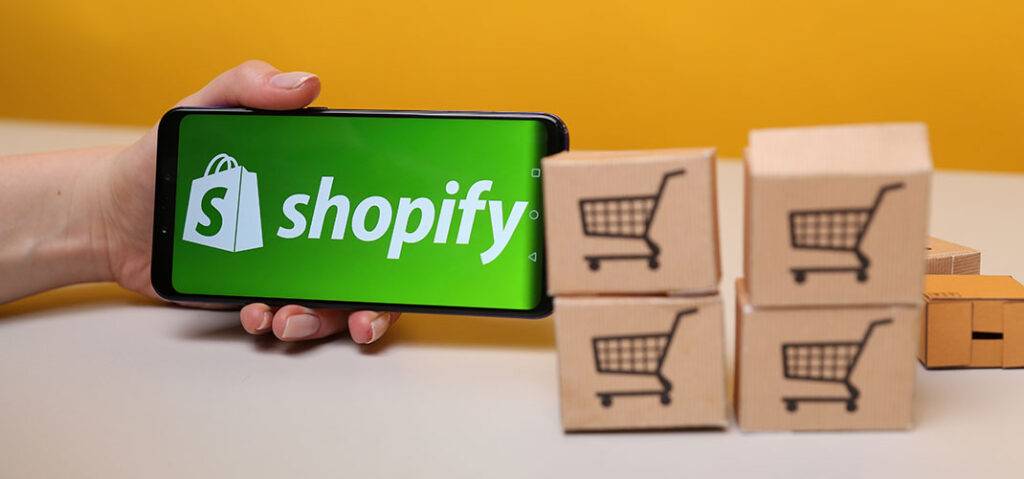 Shopify logo on iPhone beside mini cardboard boxes with shopping carts on them
